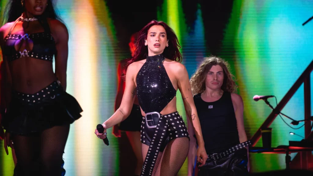 Dua Lipa Net Worth 2026: How She Built £150 Million — and Why She Is Suing Samsung for $15 Million Dua Lipa performing on stage at Glastonbury Festival 2024 in a black studded halter-neck top and studded shorts, holding a microphone, with a backing dancer and guitarist behind her against green and yellow stage lights.