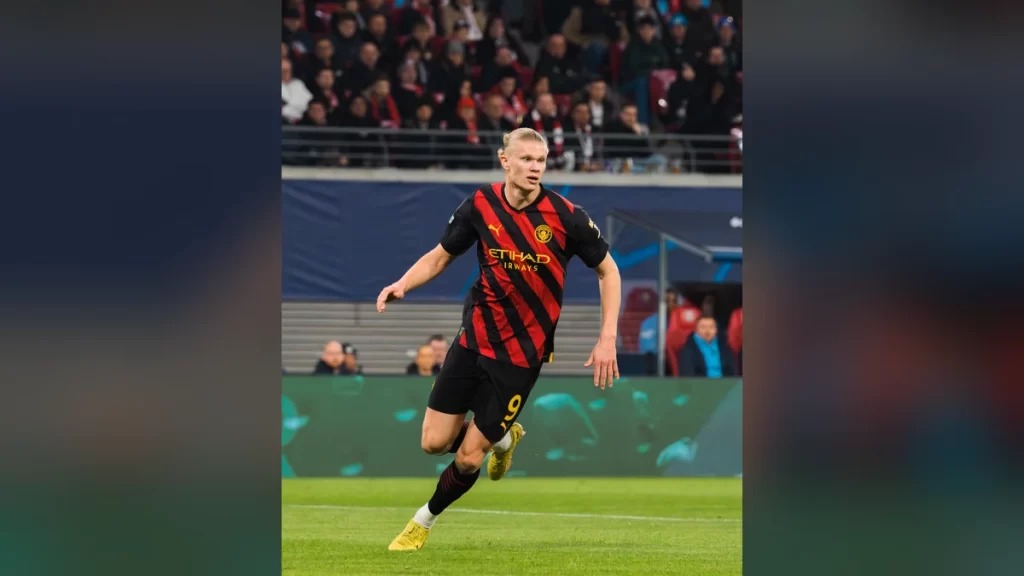 Erling Haaland running on the pitch in Manchester City's red-and-black striped away kit with the number 9 during a Champions League match at the Red Bull Arena in Leipzig, with spectators in the stands behind him.