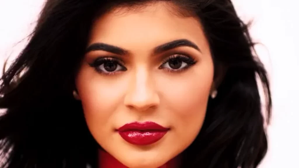 Close-up portrait of Kylie Jenner with long dark wavy hair, winged eyeliner and glossy red lipstick against a pale pink background.