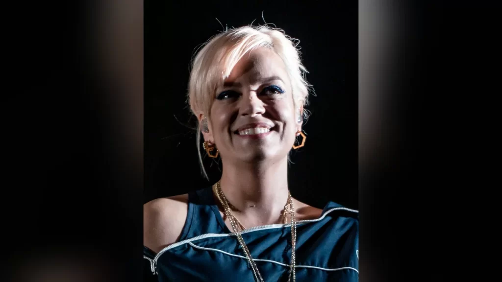 Lily Allen smiling on stage at the El Rey Theatre in Los Angeles in 2018, with short platinum-blonde hair, blue eyeliner, gold hoop earrings and layered gold necklaces, wearing a dark off-shoulder top.