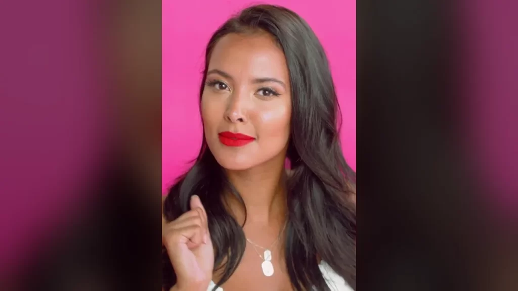 Maya Jama with long dark wavy hair and red lipstick against a bright pink background, wearing a white top and a silver pendant necklace, photographed while hosting MTV's True Love or True Lies?
