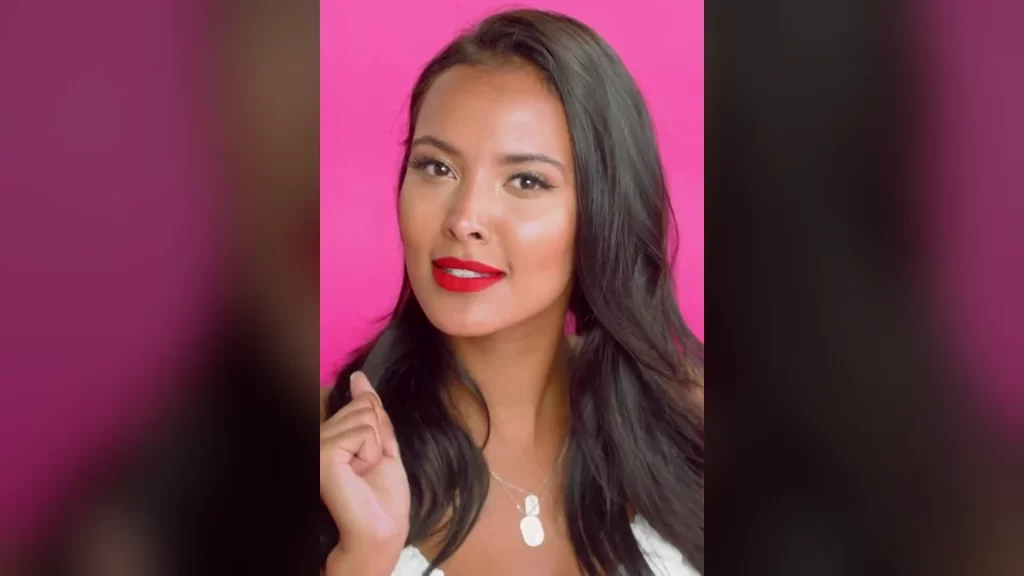 Maya Jama with long dark wavy hair and red lipstick against a bright pink background, smiling slightly with one hand raised near her hair, wearing a white top and a silver pendant necklace, photographed while hosting MTV's True Love or True Lies?