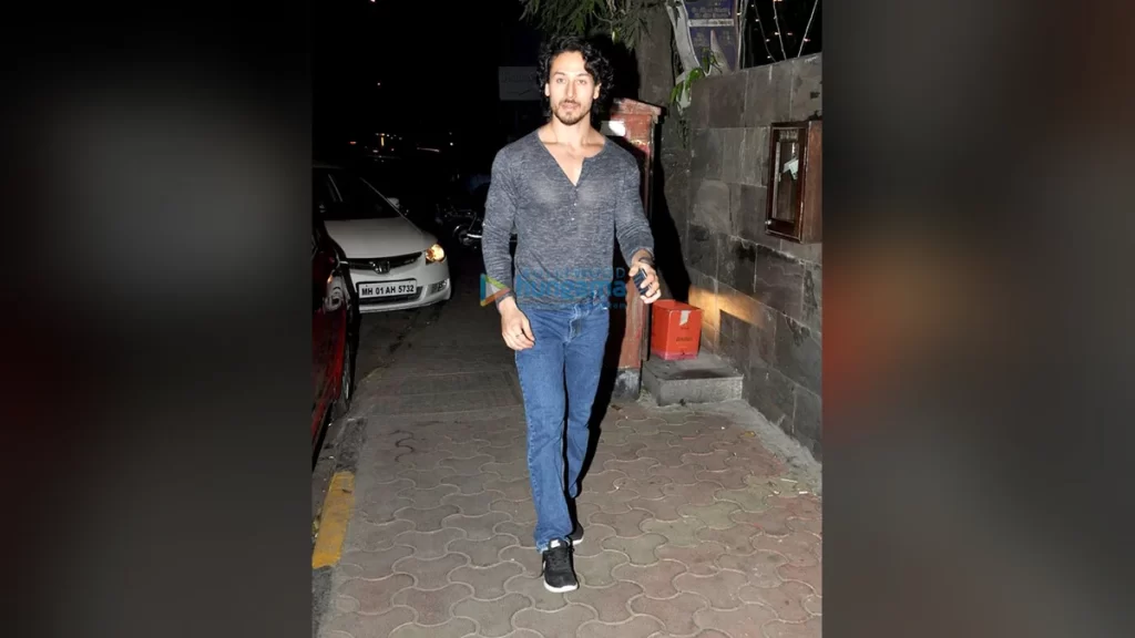 Tiger Shroff Net Worth 2026: How the Action Star Built a Rs 248 Crore Fortune Tiger Shroff walking towards the camera along a pavement in Bandra, Mumbai at night, wearing a grey long-sleeved henley shirt, blue jeans and black trainers, with parked cars behind him.